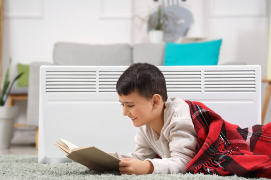 Cute boy with plaid reading book near electric convector heater on carpet at home