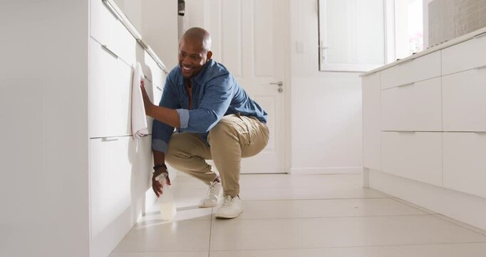 Man in blue shirt crouching at kitchen cabinets spraying with bottle and wiping cloth removing dirt
