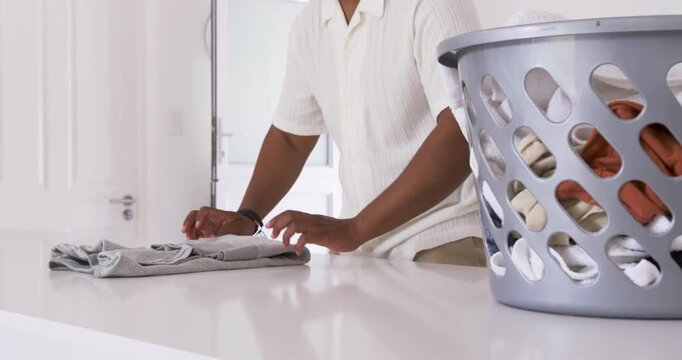 African man taking shirt from grey basket, folding and stacking clothes at counter with headphones