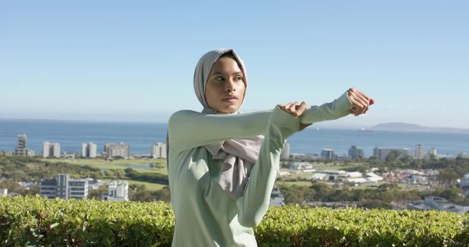 Adult female wearing hijab adjusting sleeves, stretching arms, warming up on terrace by hedge ocean