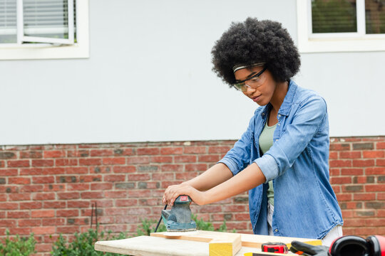 Adult African American woman using palm sander on plank at bench outside home with goggles