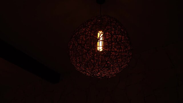 A low-light close-up of a woven spherical pendant lamp with a warm glowing bulb inside. Intricate wicker textures create a high-contrast pattern against a dark, underexposed background.
