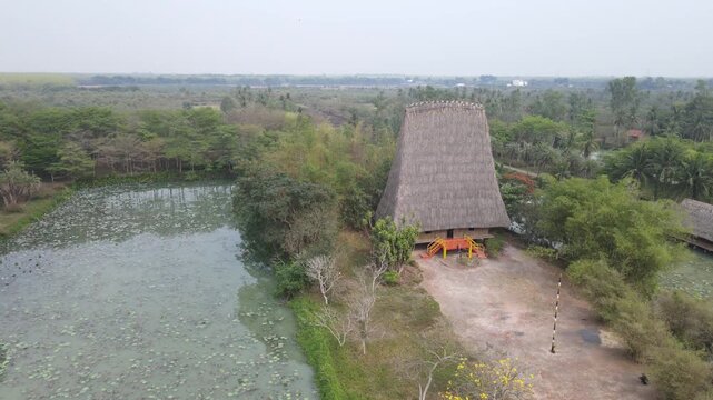 Aerial drone view of Rong House in Kon Tum showing tall thatched structure, nearby pond with lily pads, surrounding trees, and rural landscape forming cultural scene