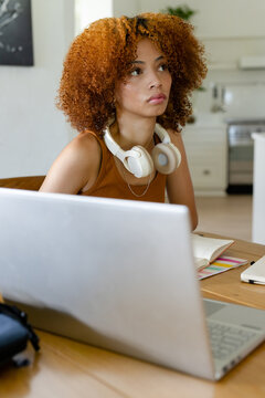 African American woman using laptop with headphones, open notebook on kitchen table in brown top