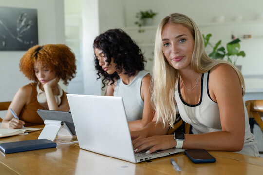 Diverse female peers collaborating at wooden table in bright kitchen using silver laptop and tablet