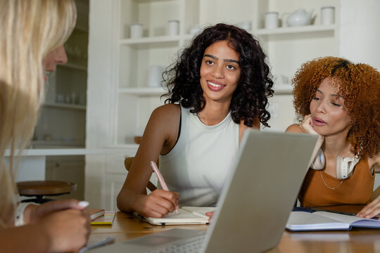 Diverse female friends studying together at home kitchen table with silver laptop and notebooks
