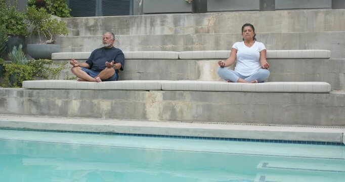 Senior African American couple meditating seated cross-legged on cushions by pool with planters