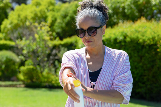 Midlife African American woman applying sunscreen to arm in garden, holding bottle with yellow cap