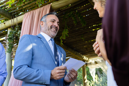 Couple standing under garden pergola exchanging vows, man holding folded papers in light blue suit