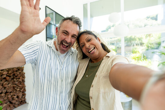 Diverse couple taking selfie in bright living room, woman holding smartphone near firewood