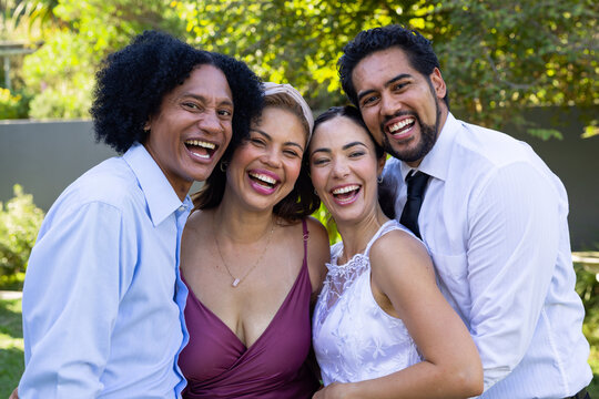 Diverse friends laughing and leaning shoulder-to-shoulder in backyard garden with dresses and tie