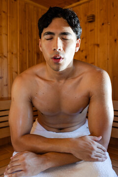 Asian man sitting on wooden sauna bench, wearing white towel, perspiring under amber light