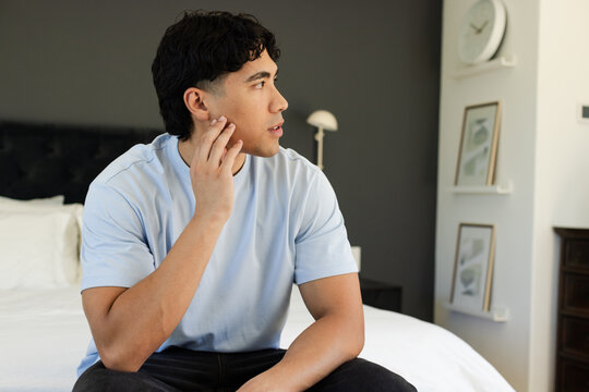Adult male sitting bedside in bedroom touching face, in blue tee, tufted headboard and clock