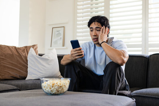 Asian man sitting on dark gray sofa holding smartphone, touching face, glass bowl of popcorn nearby
