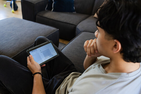 Asian man sitting on sofa in living room holding tablet, resting chin, tennis balls, copy space