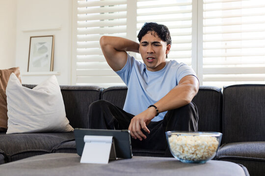 Man sitting on dark-gray sofa at home, wearing light blue T-shirt, using tablet with popcorn