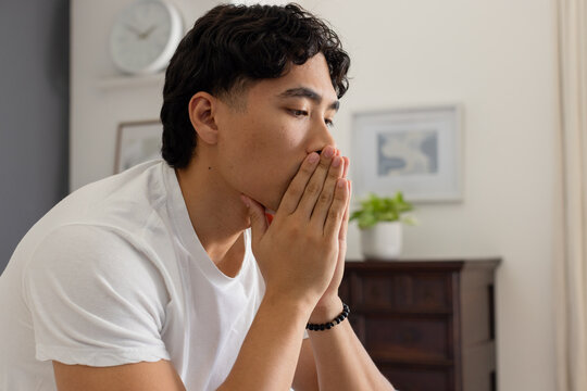 Adult Asian man sitting home covering mouth nose wearing white tee black beaded bracelet