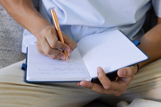 Asian adult man sitting on couch wearing white shirt writing in notebook with rose-gold pen