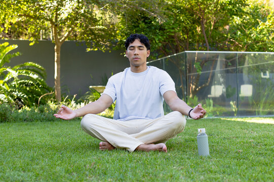 Asian man sitting cross-legged meditating in tee on garden lawn with bottle and bracelet