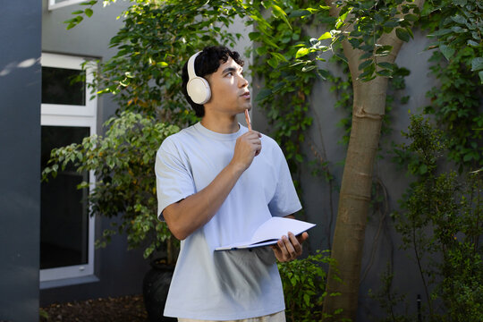Asian man standing in garden near window wearing white headphones holding open notebook and pencil