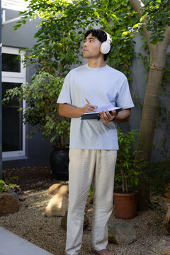 Asian man standing, listening on white headphones and writing with pencil in courtyard
