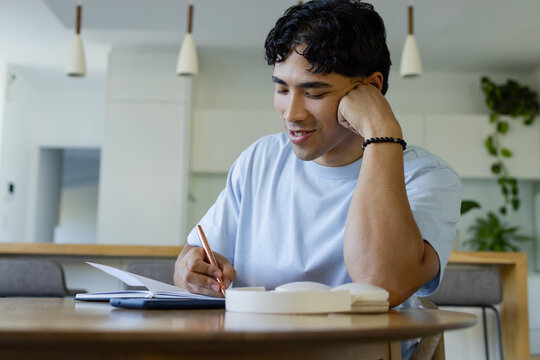 Man in early 20s writing in open notebook at modern kitchen table, holding pen, phone nearby