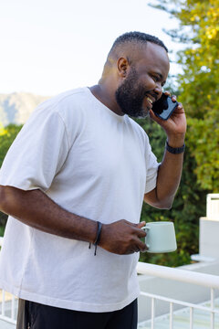 African American man wearing white T-shirt, standing on balcony holding phone and light green mug