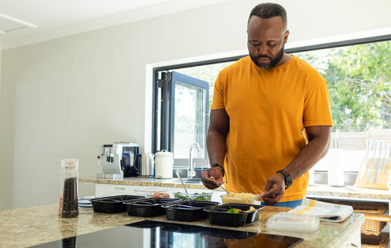 Man lining up black meal-prep containers, scooping grains from clear tub, preparing weekly lunches