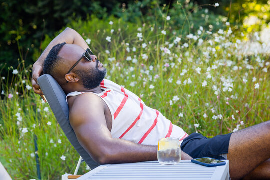 Mid-adult African man reclining on lounger beside table in backyard with smartphone, lemon glass