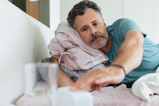 Man lying in bed reaching for alarm clock beside water glass, wearing teal tee and wristband