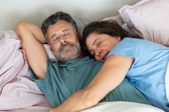 Mature couple sleeping closely in bed with pastel pillows, pale sheets and T-shirts