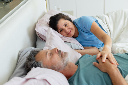 Diverse couple lying on bed at home wearing T-shirts, woman reaching across touching chest