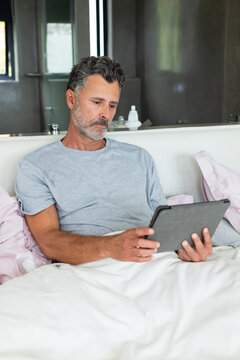 Mature man sitting against headboard in bed wearing light gray T-shirt holding tablet and reading