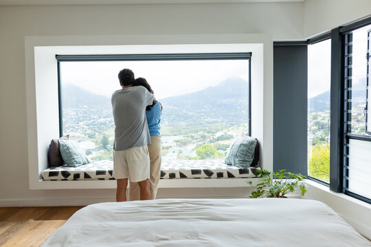 Diverse couple standing and embracing on bedroom window seat, overlooking valley with white bedding