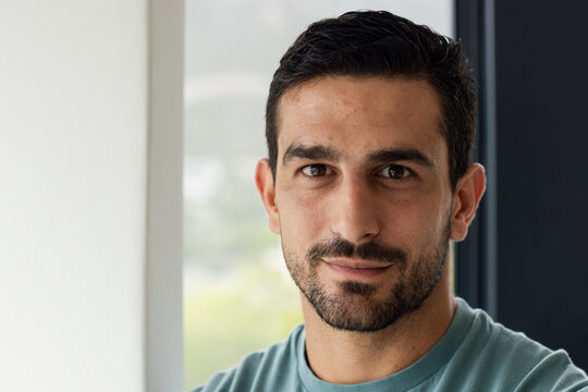 Adult male standing by window inside room, wearing teal t-shirt, trimmed beard, mullion and foliage
