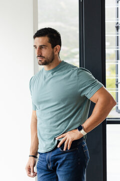 Male standing near tall window in modern room, gazing out through blinds, wearing teal shirt