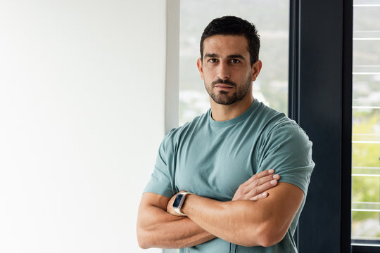 Adult male standing arms crossed facing camera by tall window wearing teal tee, smartwatch visible