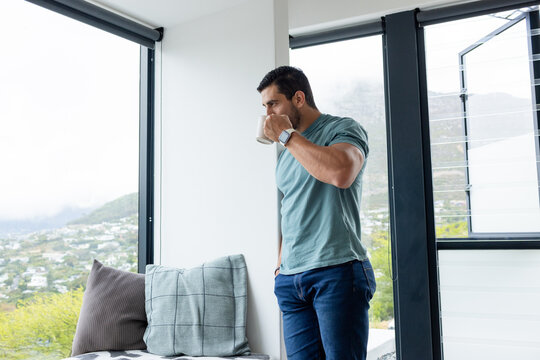 Man standing, drinking from mug by window nook in home, showing smartwatch, copy space