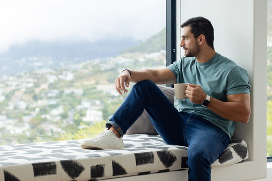 Adult man sitting on patterned window seat holding ceramic mug, smartwatch visible, copy space