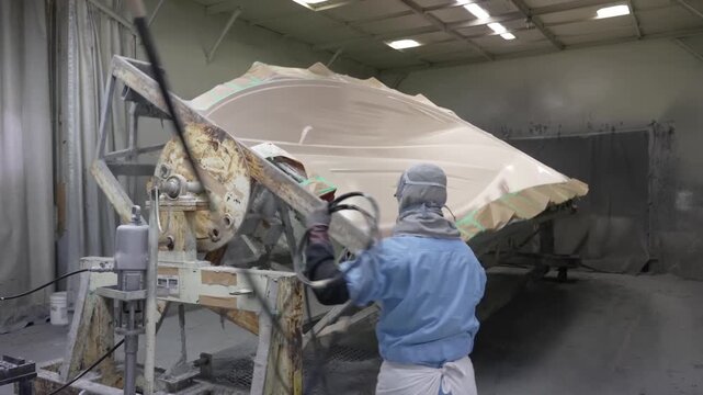 Wide shot of an industrial worker maneuvering a massive rotating metal jig holding a large boat hull in a manufacturing facility