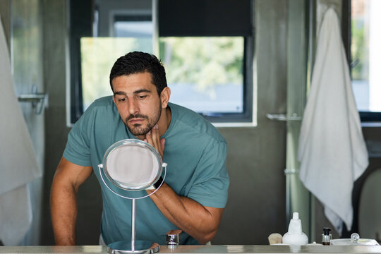 Man in teal tee leaning inspecting face touching jawline at bathroom vanity with round mirror