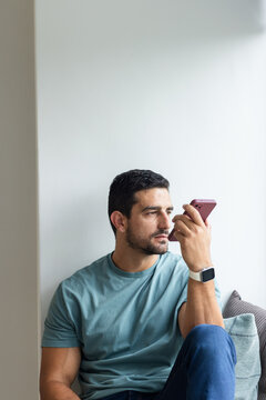 Man in his thirties sitting on couch at home holding mauve smartphone near face, wearing smartwatch