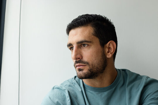 Adult man gazing left by bright window, wearing teal crewneck, leaning on backrest near blind cord