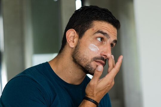 Man applying facial cream, looking into mirror in bathroom, wearing teal shirt and bracelet