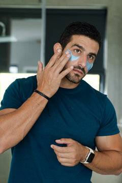 Man applying blue under-eye gel patches at bathroom mirror wearing teal t-shirt and smartwatch