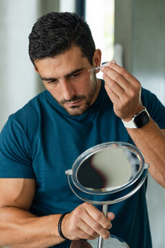 Adult man wearing teal T-shirt and smartwatch holding mirror, applying serum with dropper at vanity