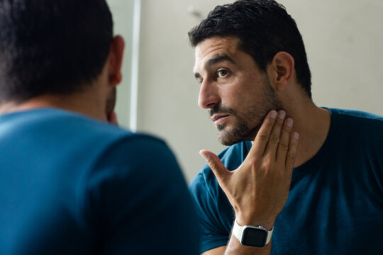 Man wearing teal tee and smartwatch touching jaw, checking face in vertical bath mirror, copy space