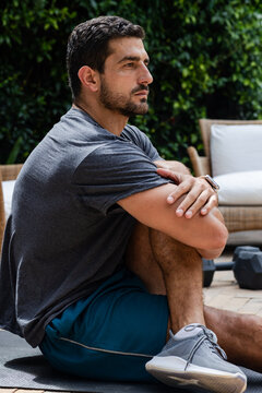 Adult man sitting on exercise mat on stone patio, wearing grey tee and fitness tracker, resting