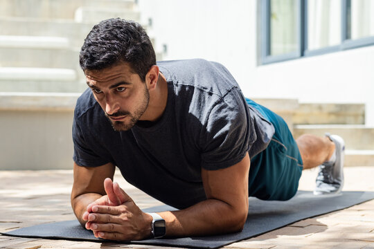 Man holding forearm plank on sunlit patio on exercise mat wearing dark grey t-shirt and smartwatch