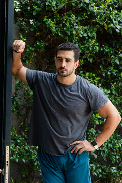 Adult man standing at doorway leaning on metal frame wearing gray tee, shorts, smartwatch, ivy wall